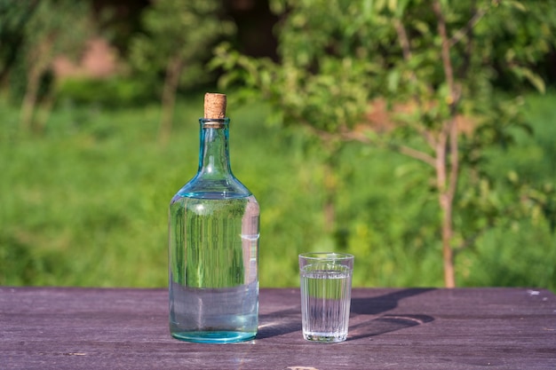 Vidrio y botella grande con agua potable en una mesa de madera sobre fondo de naturaleza en el jardín, de cerca, Ucrania. Puede ser agua o vodka