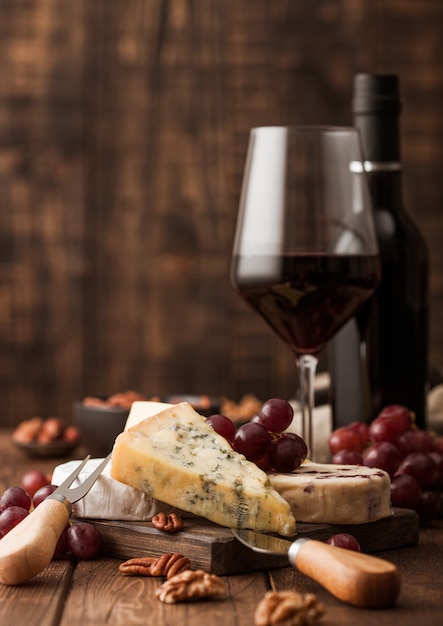 Vidrio y botella de vino tinto con selección de varios quesos en el tablero y uvas sobre fondo de mesa de madera. Blue Stilton, Red Leicester y Brie Cheese y cuchillo.