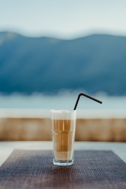 Vidrio facetado con café con leche y una pajita sobre un fondo azul, sobre una mesa en un café al aire libre. A