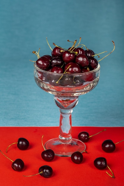 Vidro delicado cheio de cerejas maduras em um fundo azul e vermelho