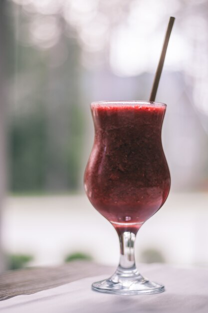 Vidro parcialmente desfocado de batido de frutas vermelhas com canudo, na mesa de madeira com toalha de mesa branca perto da janela. vertical