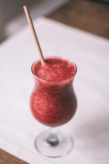 Vidro parcialmente desfocado de batido de frutas vermelhas com canudo, na mesa de madeira com toalha de mesa branca. Vertical
