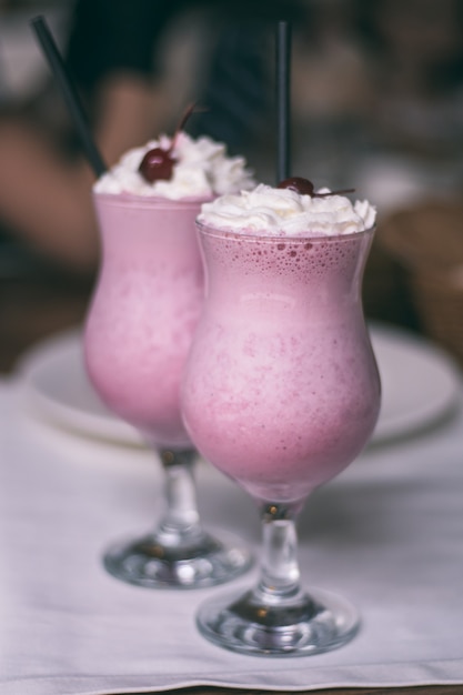 Vidros parcialmente desfocados com milkshake de baga com canudos, chantilly e coquetel de cerejas, na mesa de madeira com toalha de mesa branca. Vertical