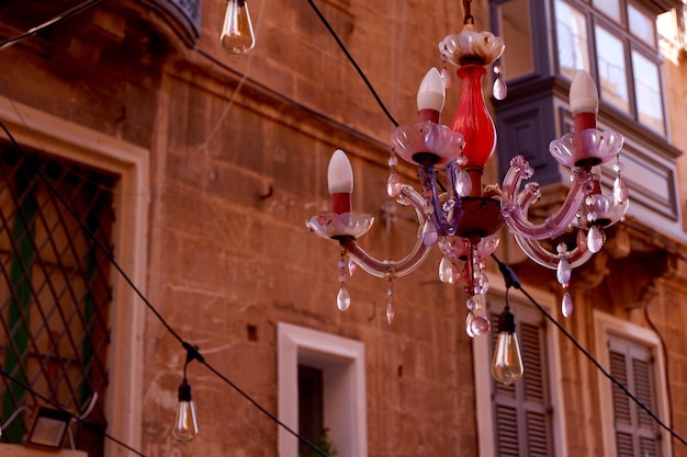 Una vieja lámpara de araña vintage en tonos rojos y rosas cuelga afuera entre casas marrones como decoración festiva Calle estrecha y hermosa de Valletta Malta