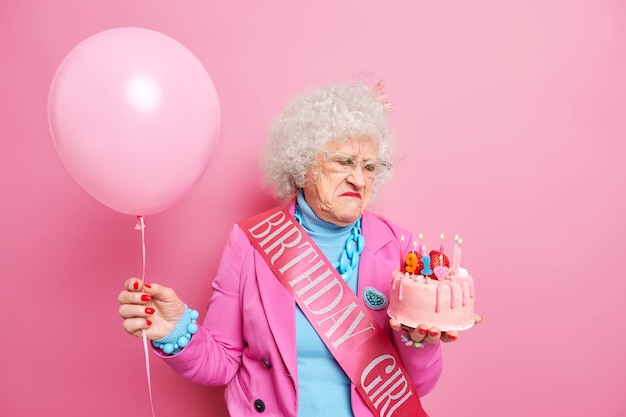 Vieja mujer hermosa de cabello gris parece disgustada con el pastel de cumpleaños, triste por envejecer, usa gafas, traje festivo, sostiene un globo inflado, acepta felicitaciones