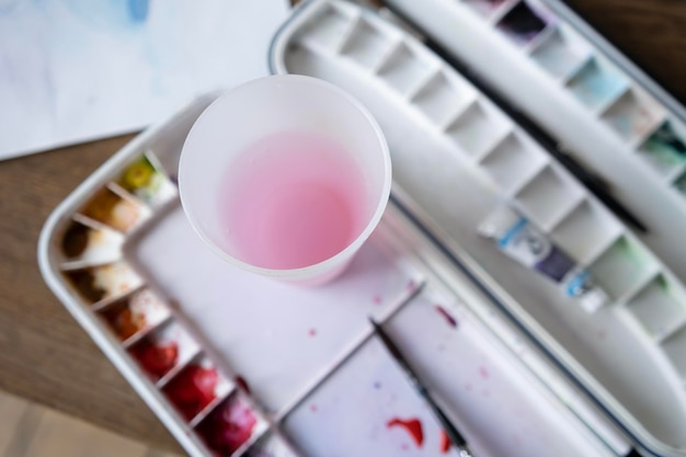 una vieja paleta con pinturas y un vaso de agua sobre una mesa de madera