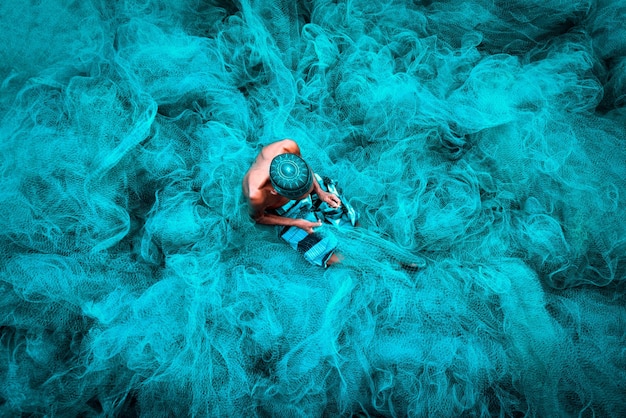 Viejas manos de pescadores cosiendo redes de pesca azules sentadas en el suelo y rodeadas de una gran red