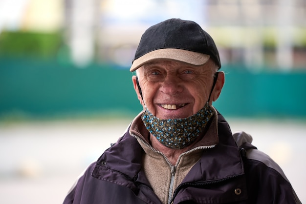 El viejo abuelo, con una máscara médica, una gorra, sonríe y mira a la cámara.