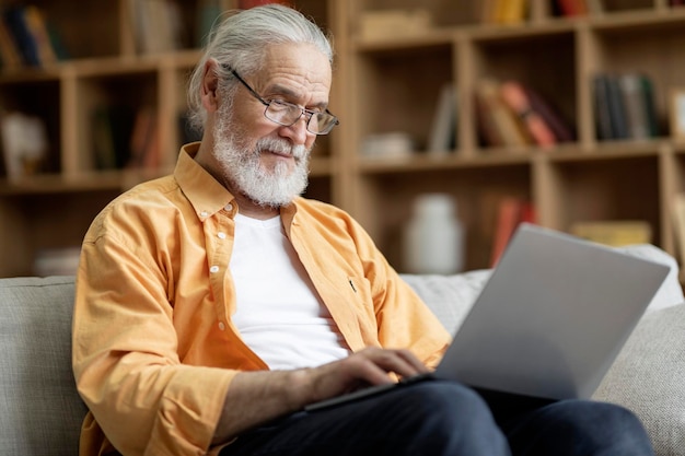 Viejo en anteojos usando una computadora portátil moderna en casa