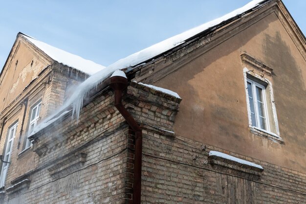 Viejo edificio de ladrillo cubierto de nieve de fuertes nevadas Colgando carámbanos del techo de un edificio residencial Vista del edificio de ladrillo rojo contra el cielo azul peligro de caída de carámbanos