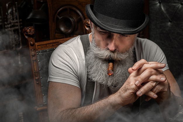 Viejo fumando un cigarro en el interior. Concepto de la mafia. Elegante hombre en su gabinete. Hombre barbudo.