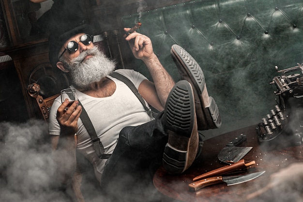 Viejo fumando un cigarro en el interior. Concepto de la mafia. Elegante hombre en su gabinete. Hombre barbudo.