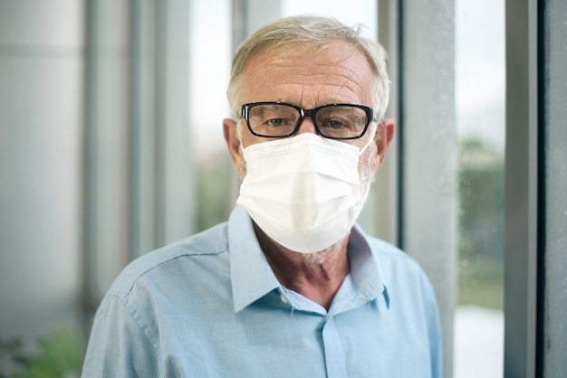 Viejo hombre caucásico lleva máscara médica, mirando hacia fuera de su casa