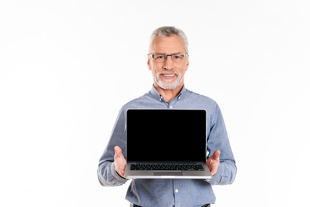 Viejo hombre sonriente que muestra la computadora portátil con la pantalla en blanco aislada