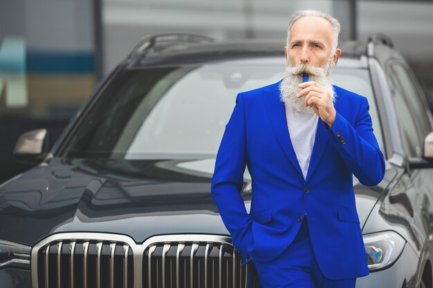 Viejo hombre styish de pie cerca del automóvil de lujo. Hombre rico maduro en el traje al aire libre cerca del coche.