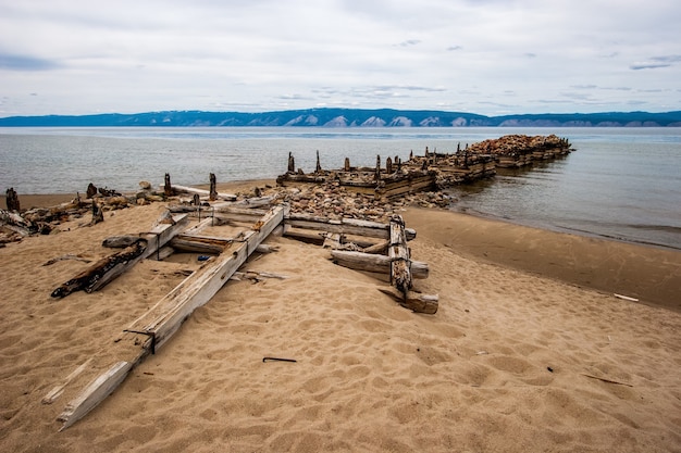 Viejo muelle del barco roto en la isla de Olkhon en el lago Baikal. Barras y troncos podridos. En la orilla de piedras y arena. Detrás del lago de montaña. El clima es lúgubre, el cielo gris.