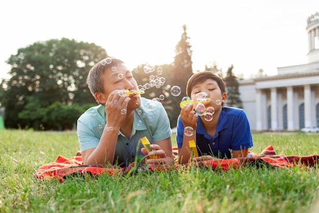 El viejo padre asiático junto con su hijo se acuestan en la hierba en el parque y soplan burbujas de jabón y se regocijan