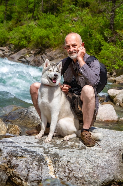 Foto el viejo y el perro de trineo caminan cerca del río. paisaje alpino. pensionista de ocio activo. anciano está sonriendo. camina con el husky siberiano.