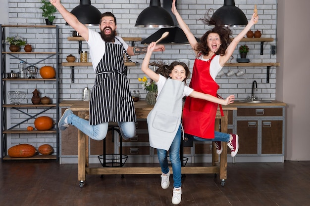 Viel Spaß in der Küche. Familie Mama Papa und kleine Tochter tragen Schürzen in der Küche springen. Familie, die Spaß beim gemeinsamen Kochen hat. Bringen Sie dem Kind bei, Essen zu kochen. Frühstück am Wochenende. Kochen mit Kind kann Spaß machen.