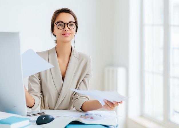 Vielbeschäftigte erfolgreiche Managerin arbeitet mit Papieren im Büro, posiert am Schreibtisch, trägt Brille und formelles Outfit, bereitet den Bericht vor, sieht selbstbewusst aus