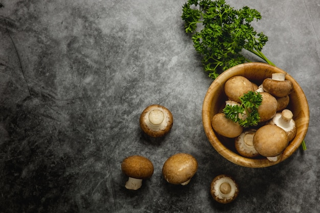 Viele braune Champignons in einer Holzschale auf dem schwarzen Zementboden Pilze fallen auf den Boden Mit einer Petersilie daneben. Gesundes Lebensmittelkonzept und vegetarisches Essen. Dunkler Ton. Draufsicht und Kopierraum