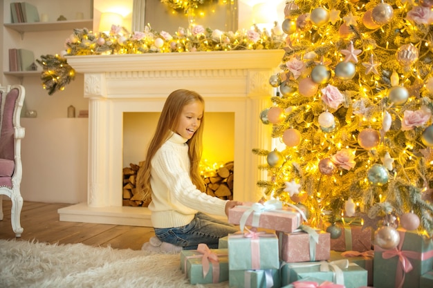 Viele Geschenke. Blondes süßes Mädchen, das beschäftigt schaut und die Weihnachtsgeschenke betrachtet