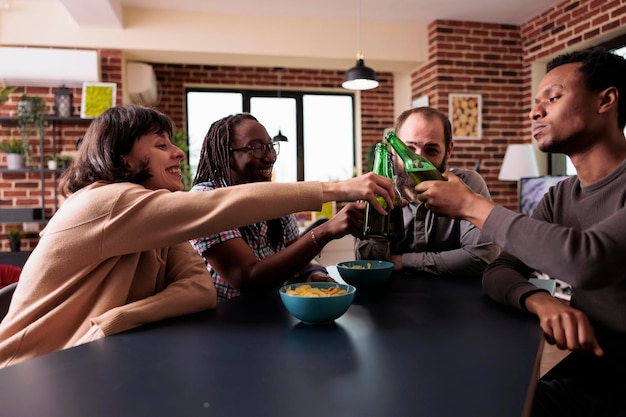 Vielfältige Gruppe von Freunden, die zu Hause ein Wiedersehen mit Snacks und Bier feiern. Fröhliche positive junge Erwachsene, die Zeit miteinander genießen, während sie alkoholische Getränke im Wohnzimmer trinken.