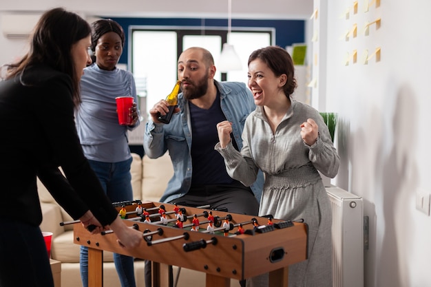 Vielfältiges Team von Kollegen, die nach der Arbeit Tischfußball-Fußballspiel spielen. Multiethnische Gruppe, die lustige, fröhliche Aktivitäten genießt, während sie im Büro Bieralkohol aus Tassenflaschen trinkt