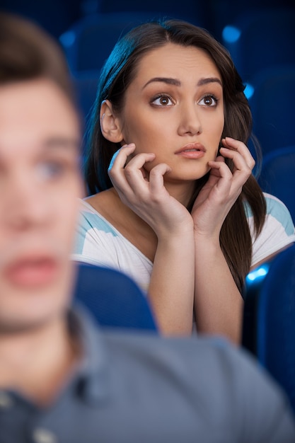 Viendo una película emocionante. Mujer joven emocionada que lleva a cabo las manos en la barbilla mientras ve la película en el cine