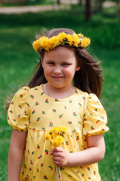 El viento sopla el cabello de una niña con un vestido amarillo y una corona de dientes de león amarillos en la cabeza en ...