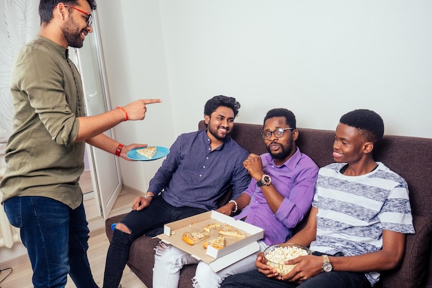 Vier afroamerikanische Männer essen Pizza auf einer Party zu Hause und werfen Popcorn ineinander.