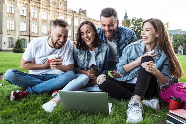 Vier beste junge Studentenfreunde mit einem Laptop auf dem Rasen im Universitätsgebiet sind glücklich und aufgeregt