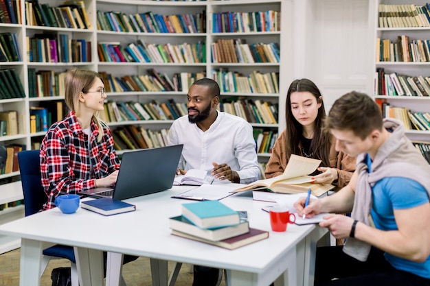 Vier gemischtrassige glückliche Studenten sitzen am Tisch in der Bibliothek, während sie lernen und an einem Laptop arbeiten.