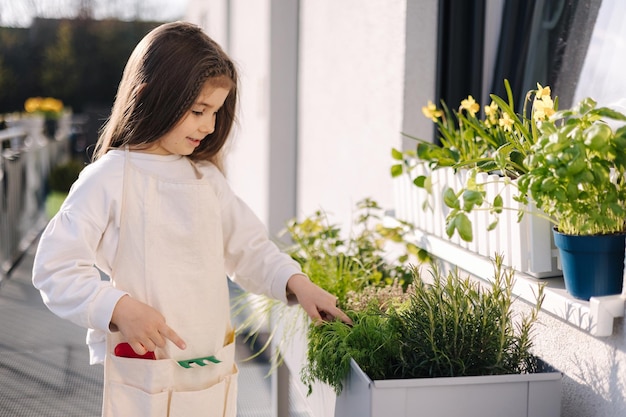 Vier Jahre altes Mädchen in hellem Overall steht am Blumentopf und bereitet sich auf die Landung auf dem Balkon vor