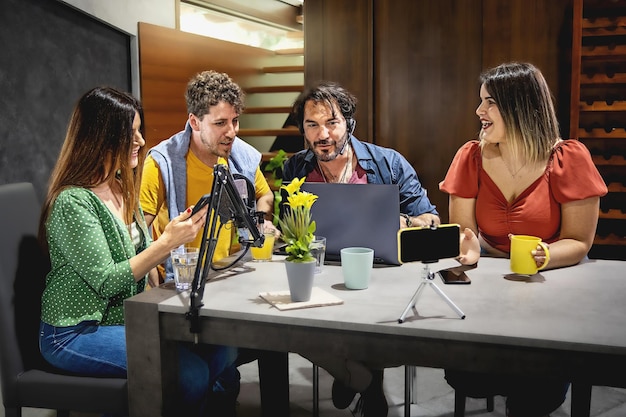 Vier junge Influencer amüsieren sich beim Streamen eines Vlog-Video-Podcasts, sitzen am Studiotisch mit Mikrofon und nehmen mit einem Handy auf einem kleinen Stativ auf
