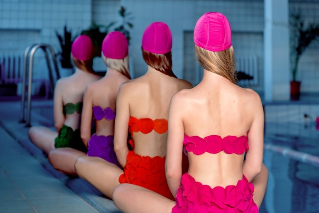 vier schlanke junge stylische Frauen in bunten Badeanzügen und rosa Bademützen im Schwimmbad