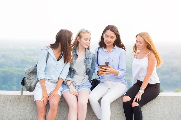 Vier schöne Frauen sitzen im Park und schauen auf das Smartphone und lächeln. Moderne Technologie, Sommer, Lifestyle-Konzepte