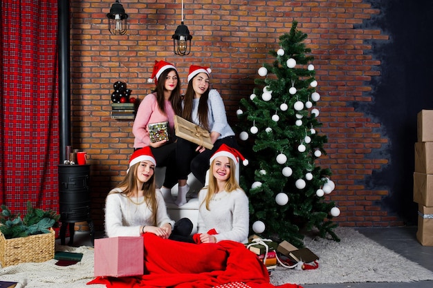 Vier süße Freundinnen tragen warme Pullover, schwarze Hosen und Weihnachtsmützen gegen den Neujahrsbaum mit Weihnachtsdekoration im Studio