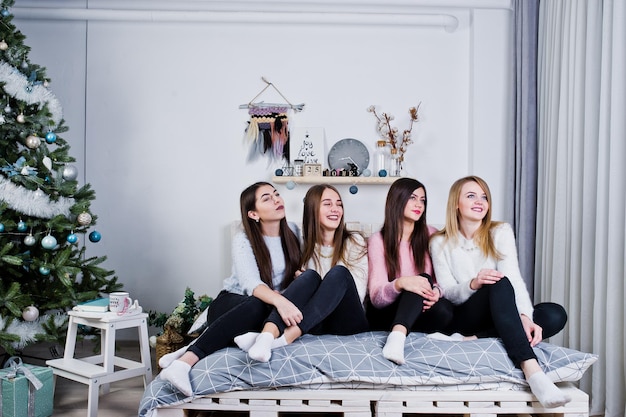 Vier süße Freundinnen tragen warme Pullover und schwarze Hosen auf dem Bett im neujahrsdekorierten Zimmer im Studio
