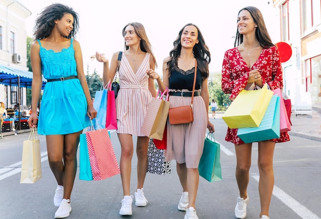 Vier wunderschöne Frauen in sommerlichen Outfits posieren auf der Straße in der Innenstadt, lächeln vor Freude und zeigen ihre Einkäufe in die Kamera