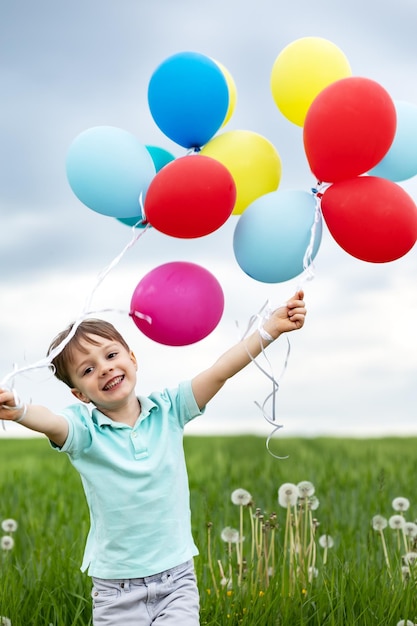 Vierjähriger Junge mit Luftballons geht auf einer grünen Wiese mit Löwenzahn spazieren