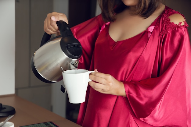 Vierta el té de cristal de agua caliente, la mujer prepara el té de la mañana para desayunar en casa, una niña en camisón de satén y una bata roja prepara el té en la cocina