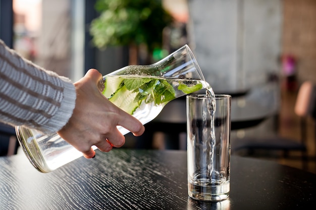 Se vierte agua con menta y pepino en un vaso de una jarra transparente sobre la mesa