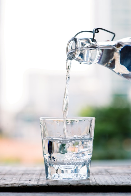 Se vierte agua potable limpia de una jarra en un vaso de vidrio redondo sobre una mesa de madera y una macro de primer plano de una servilleta verde claro sobre un fondo verde de la naturaleza al aire libre.