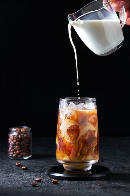 Se vierte la leche en café helado en un vaso alto sobre un fondo oscuro. Concepto Bebida Refrescante De Verano.
