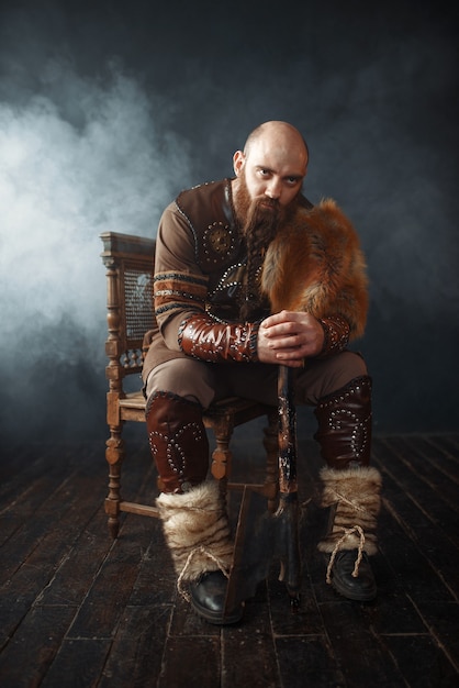 Viking barbudo com machado vestido com roupas nórdicas tradicionais sentado na cadeira