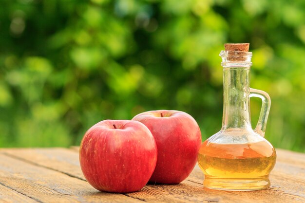 Vinagre de manzana en botella de vidrio con corcho y manzanas rojas frescas sobre viejas tablas de madera con fondo natural verde borroso