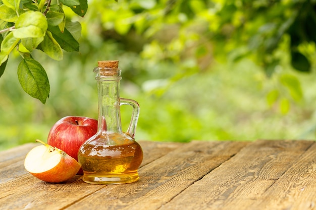 Vinagre de manzana en botella de vidrio con corcho y manzanas rojas frescas sobre viejas tablas de madera con fondo natural verde borroso