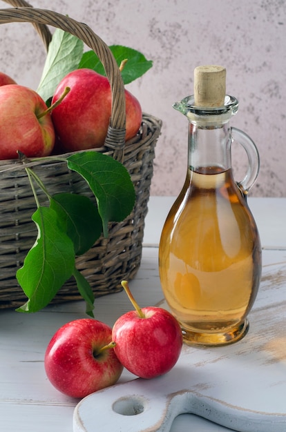 Vinagre de sidra de manzana en botella de vidrio y manzanas maduras en canasta sobre mesa de madera blanca. Alimentos orgánicos saludables.