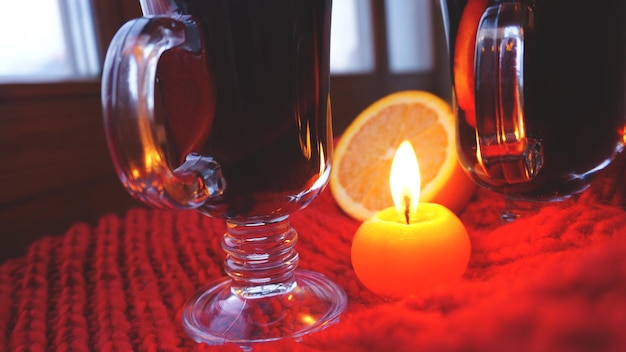 Vinho quente em canecas de vidro queimando vela sobre fundo vermelho escuro vinho quente vermelho com lenço laranja e vermelho